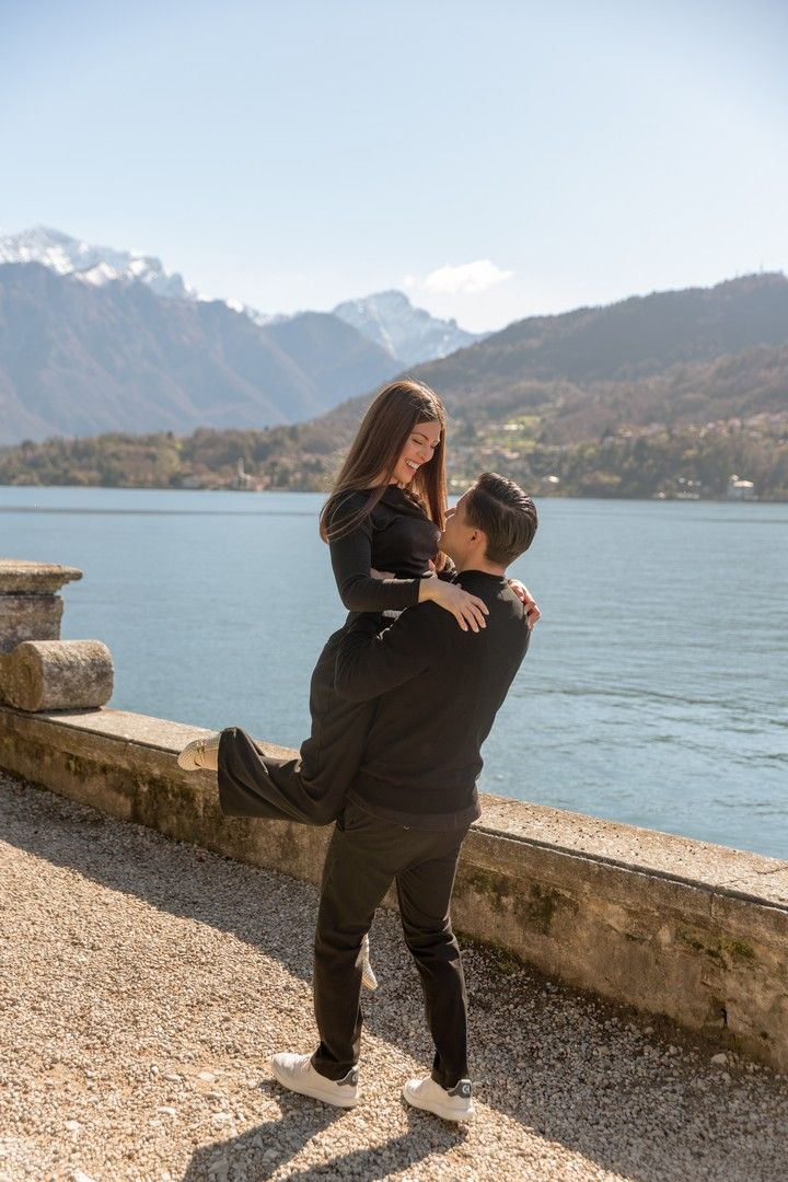 Couple in black outfits sharing a romantic lift embrace by a mountain lake with snow-capped Alps in the background.