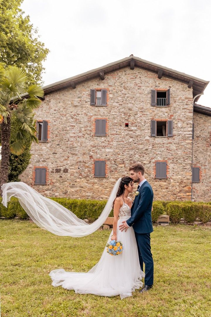 Newlyweds pose in front of a rustic stone building as the bride's veil flows in the wind.
