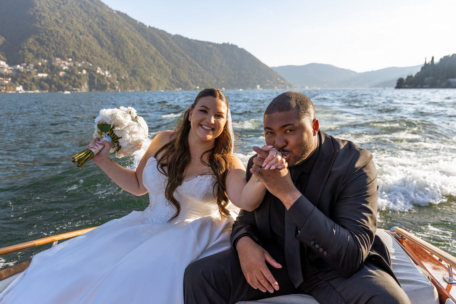 A couple shares tender moments on a boat with scenic mountain views and water in the background.