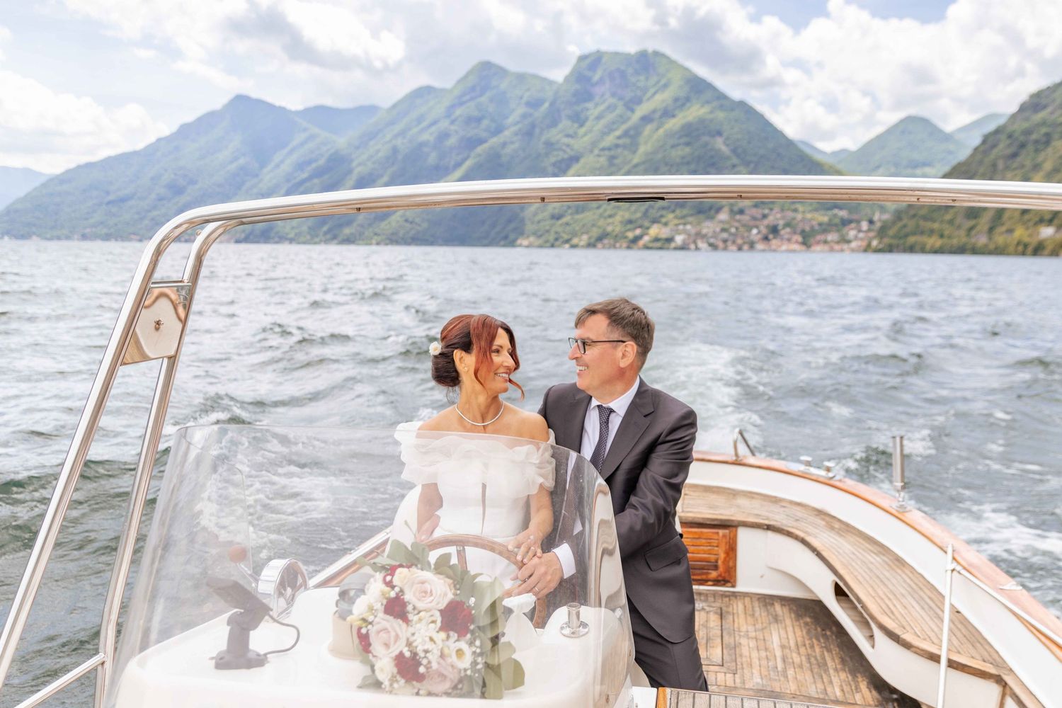 Wedding couple on classic wooden boat against scenic mountain lake backdrop.