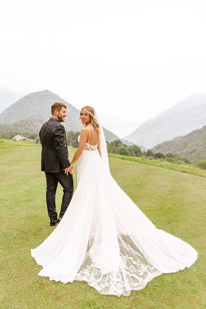 A couple in wedding attire walks together on a green hillside with mountains in the background.