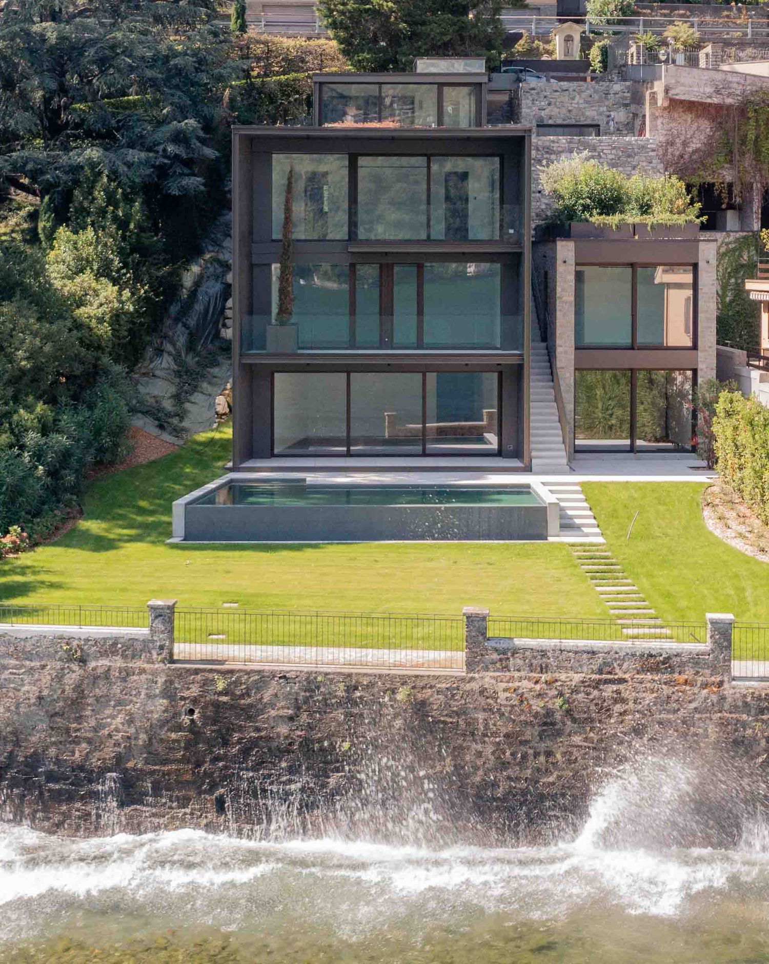 Contemporary waterfront home with waves crashing against stone wall and manicured lawn.