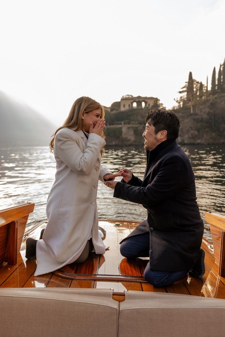 Man proposes on a wooden boat at Lake Como, Italy, with woman covering her mouth in surprise.