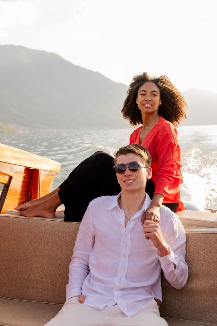 Couple relaxing on a luxury boat with mountain lake views, woman in red top and man in sunglasses holding hands.