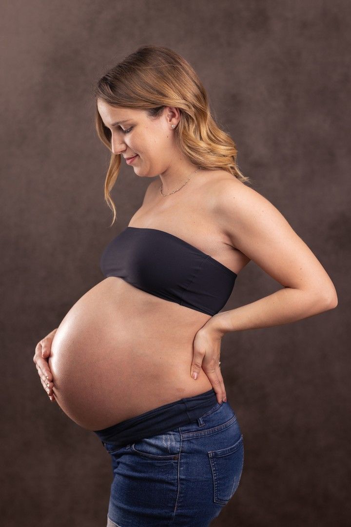 Maternity portrait of an expectant mother in black top and jeans against dark background.