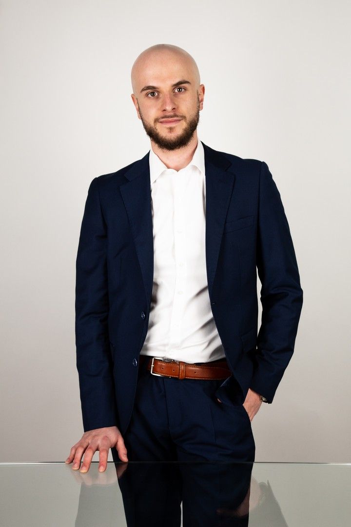 Professional portrait of person in navy suit and white shirt against light background.