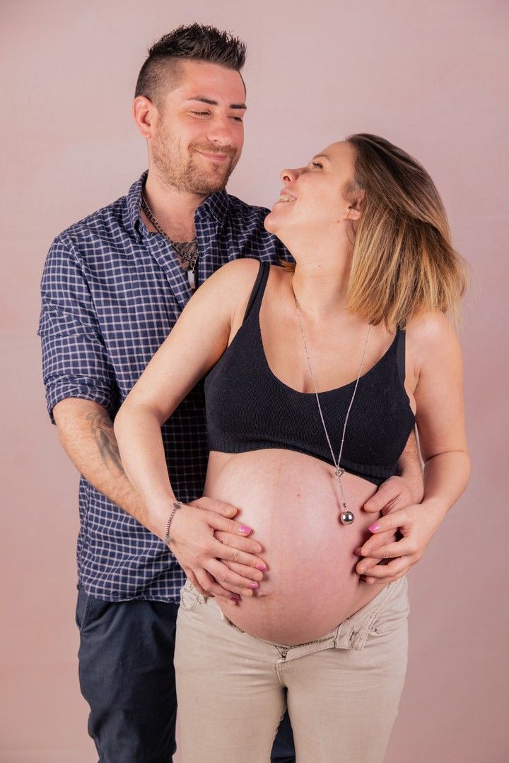 Loving couple embraces during maternity photoshoot against pink background.