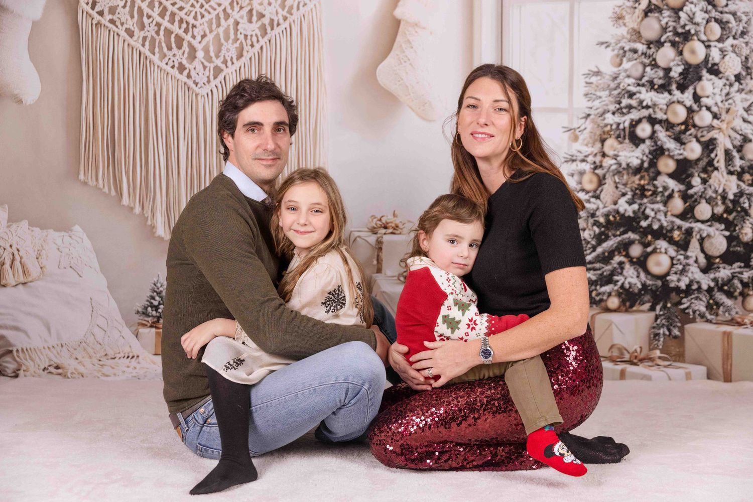 Family posing for holiday photos on white floor with decorated Christmas tree in background.
