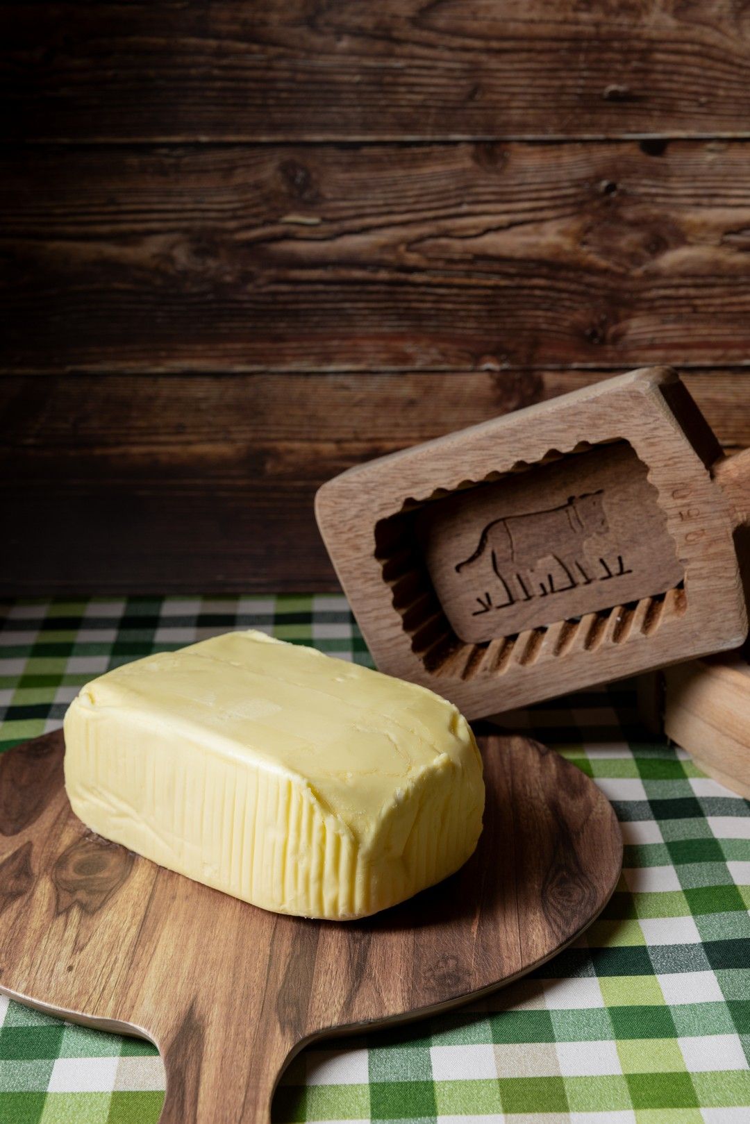 Wooden butter mold and fresh butter block on checkered tablecloth with rustic background.