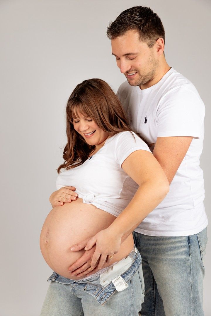 Smiling couple in white shirts cradling pregnant belly together in a studio photo.