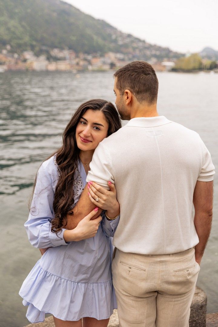Couple embracing by a lakeside waterfront, woman in blue dress smiling, scenic Italian town in background.