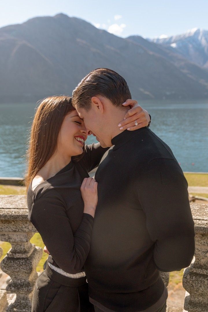 Romantic couple in matching black outfits embracing by a mountain lake with snow-capped peaks in the background.