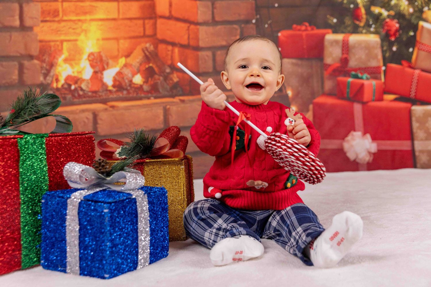 Child sits among colorful wrapped presents near brick wall and Christmas tree decorations.