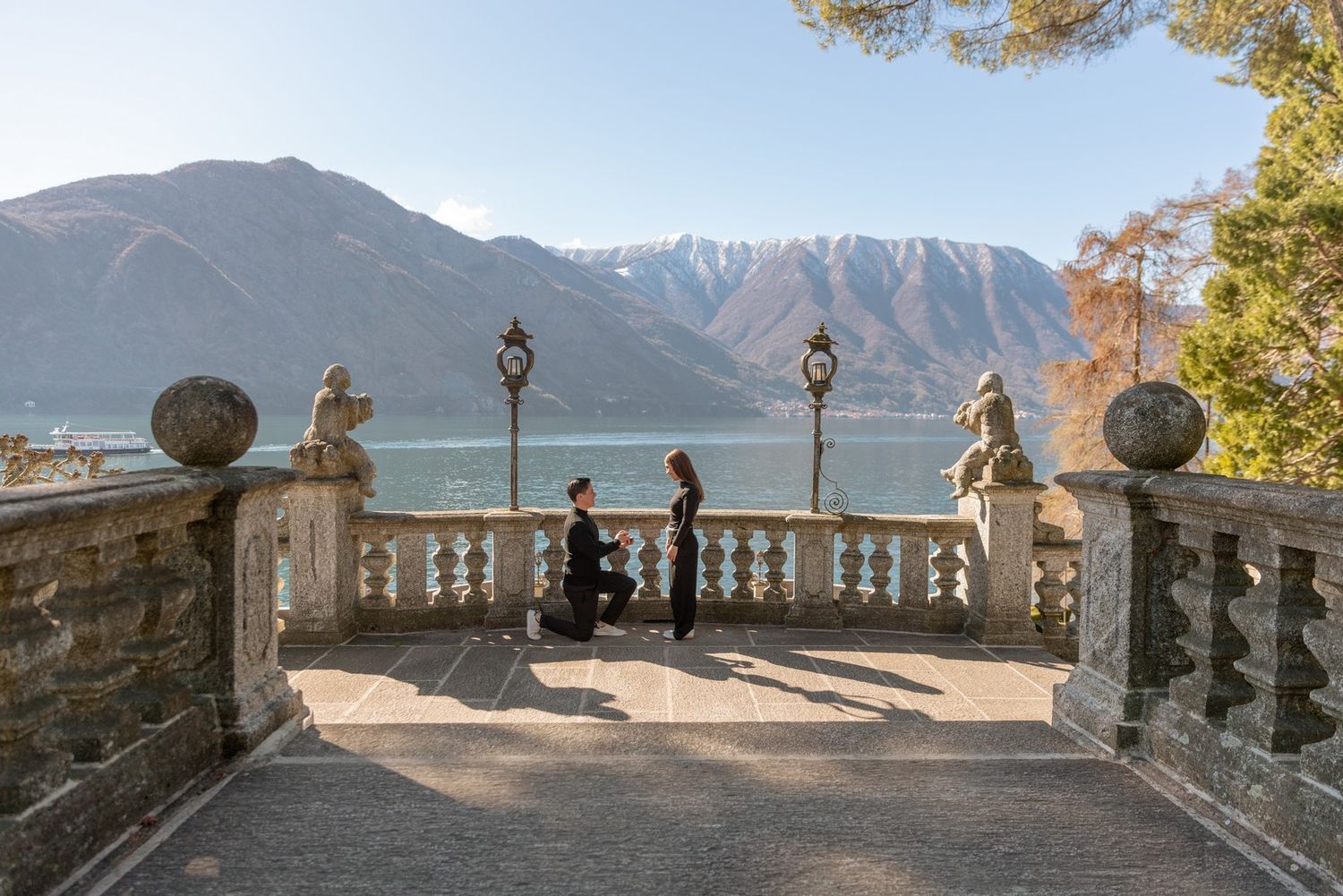 Man proposes to woman on elegant stone terrace overlooking Lake Como with mountain backdrop on sunny day.