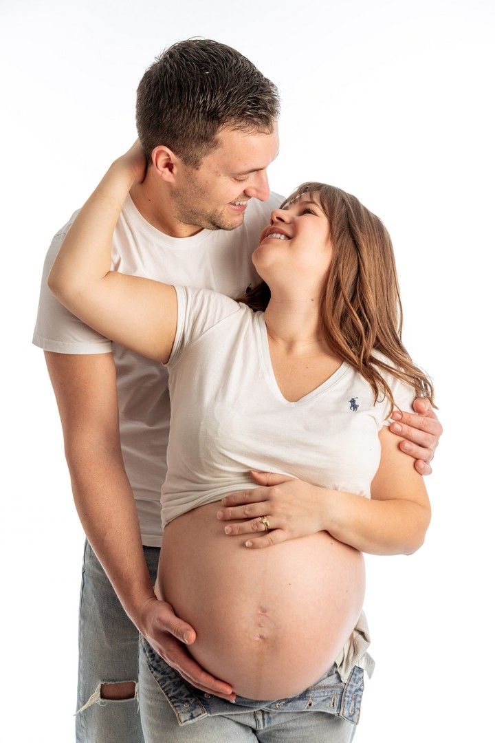 Happy couple expecting a baby, man embracing pregnant woman showing bare belly, wearing white shirts on white background.