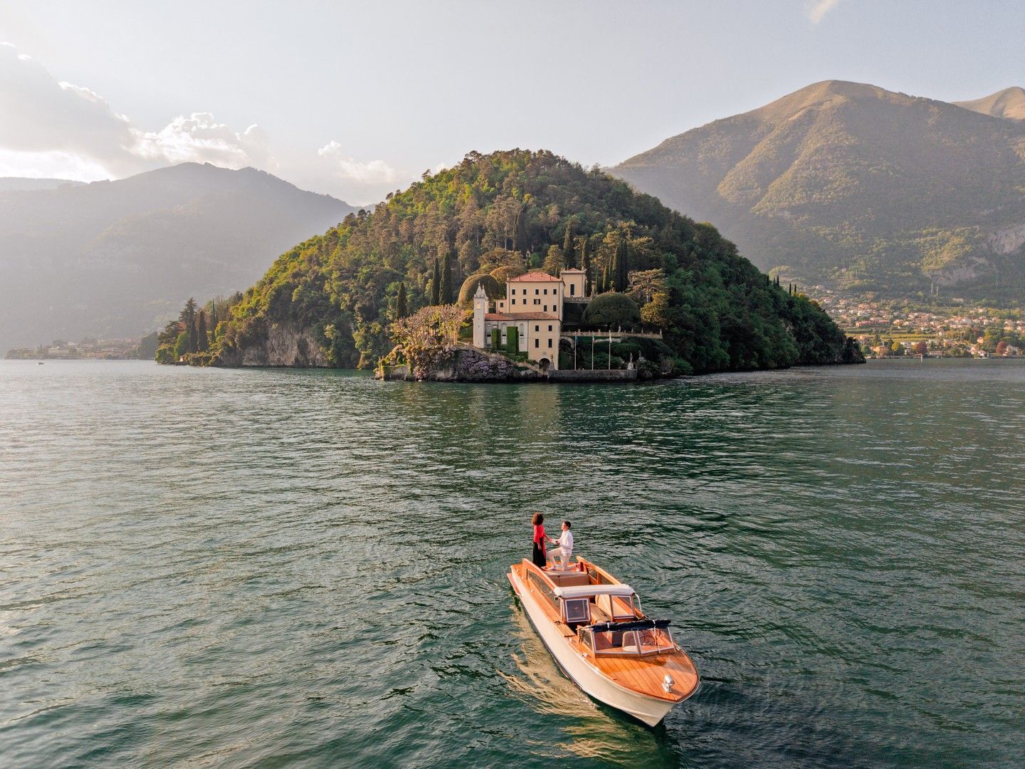 Aerial view of a classic wooden speedboat on Lake Como with a lush peninsula villa and mountains in the background.