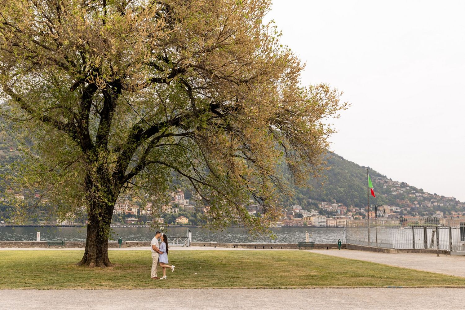 Person walking under a large tree by Lake Como waterfront with Italian flag and mountain backdrop.
