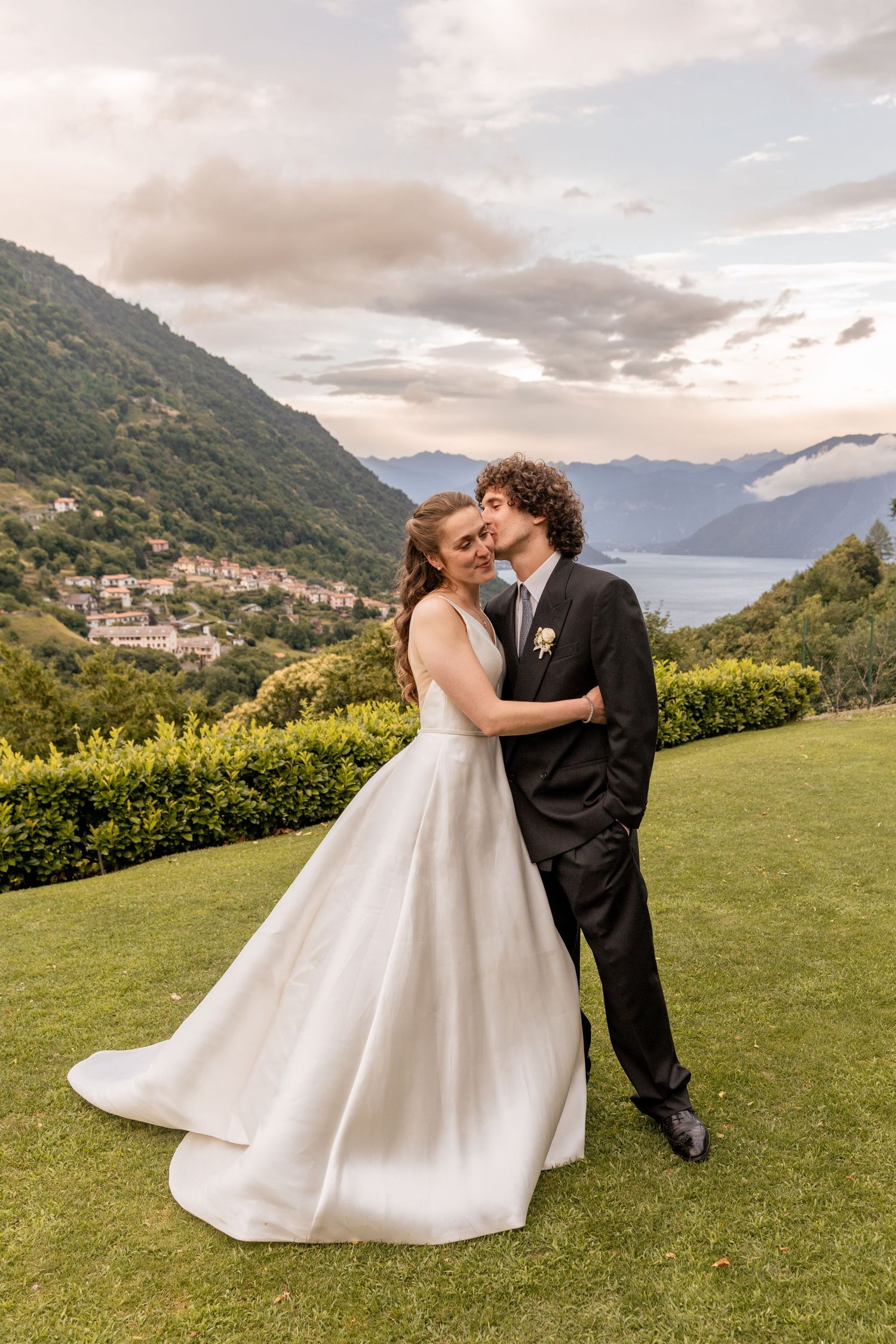 Newlyweds sharing romantic moment on hillside overlooking mountain lake vista.