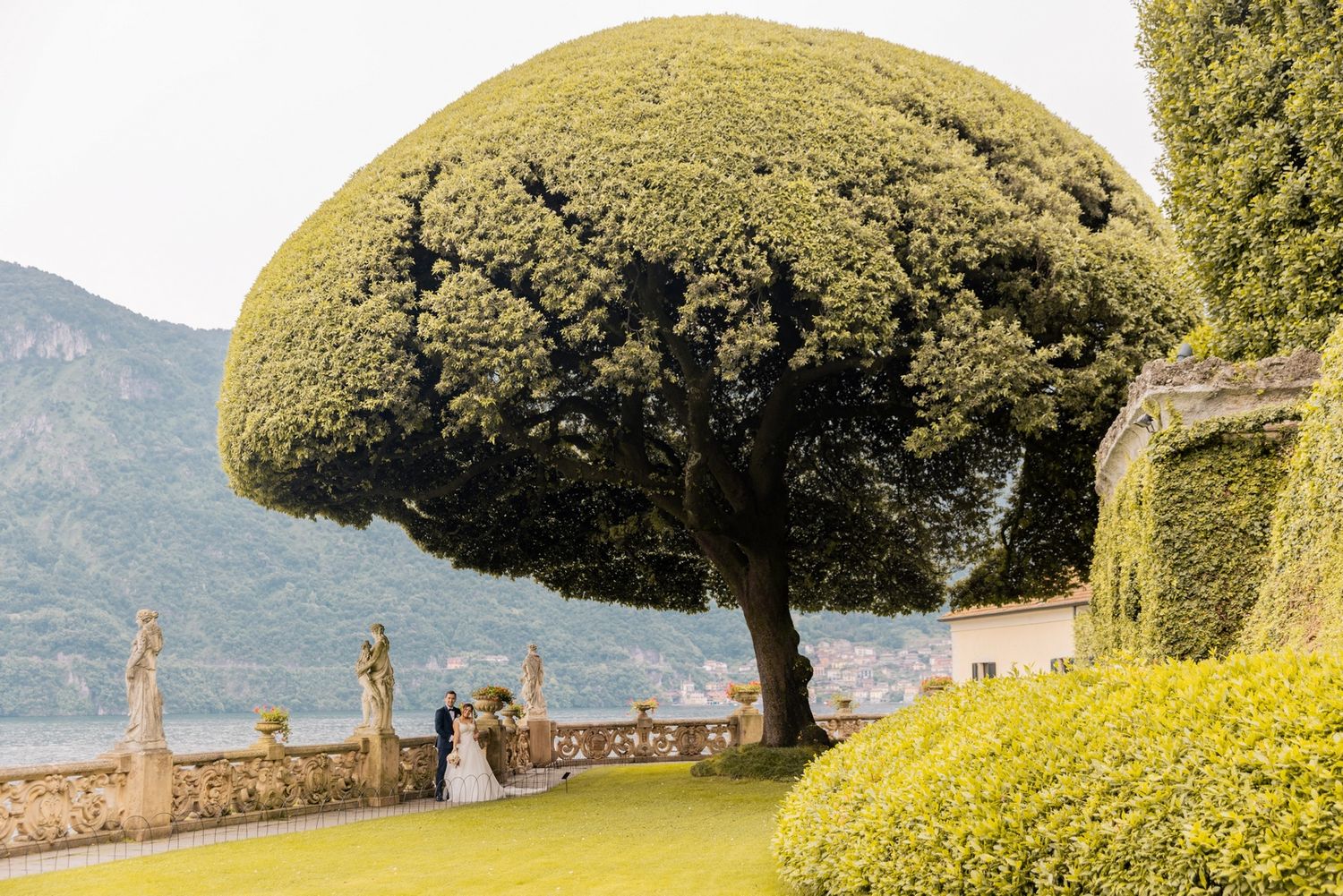 Panoramic view of Lake Como in Italy with lush gardens, stone walls and cypress trees overlooking the water.