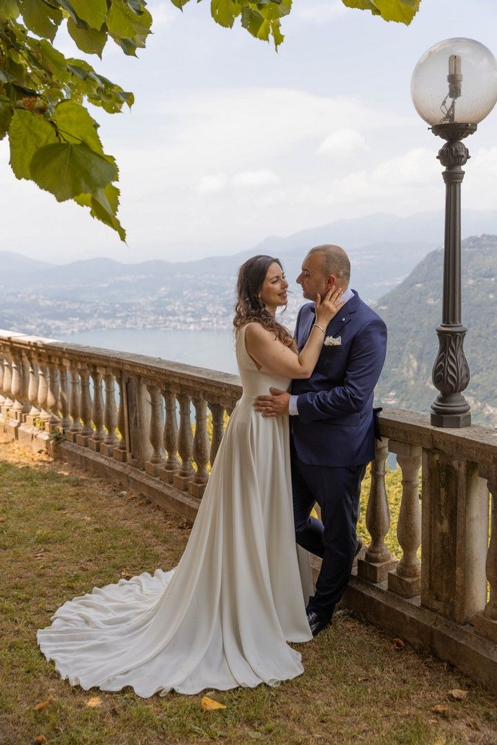 Elegant wedding couple embracing on stone balcony with lake and mountains in background.