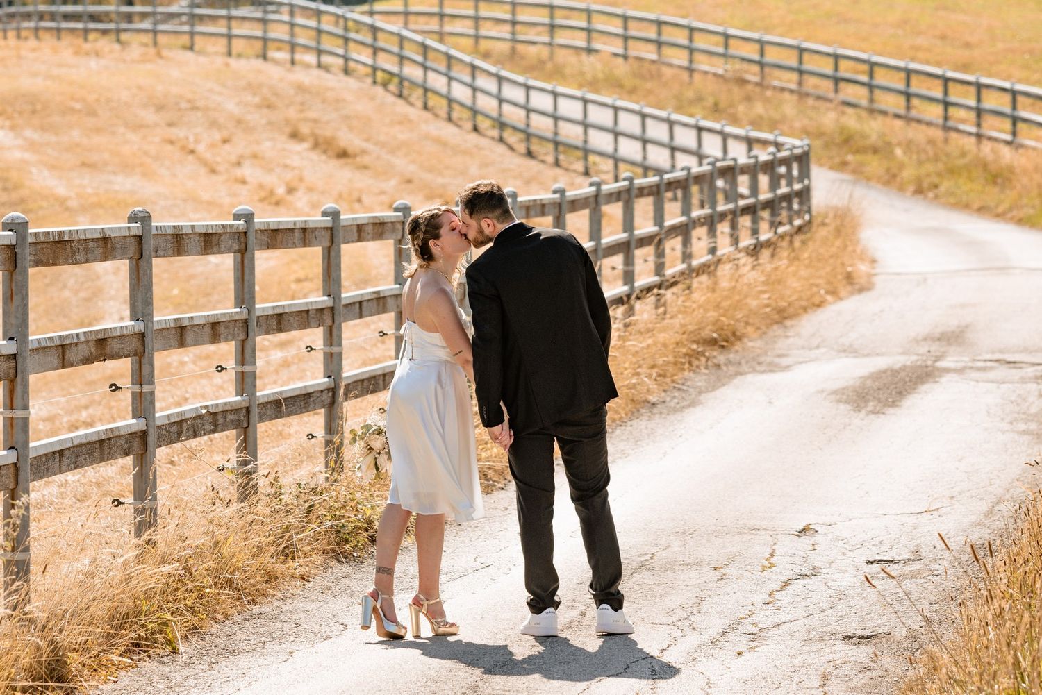 A couple walks together on a dirt path alongside a wooden fence on a sunny autumn day.