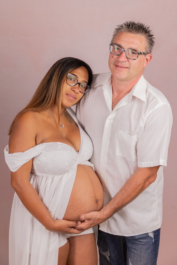 Intimate maternity photoshoot with couple in white clothing against pink background.
