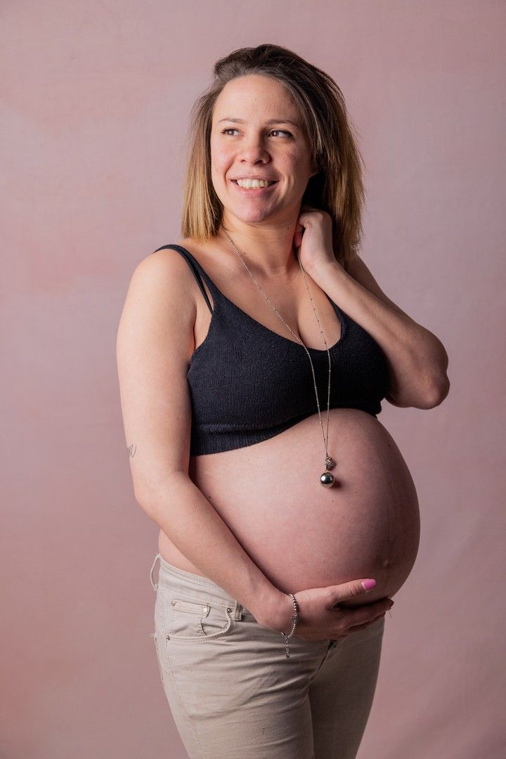 Expectant mother in black top poses against pink background while holding belly.