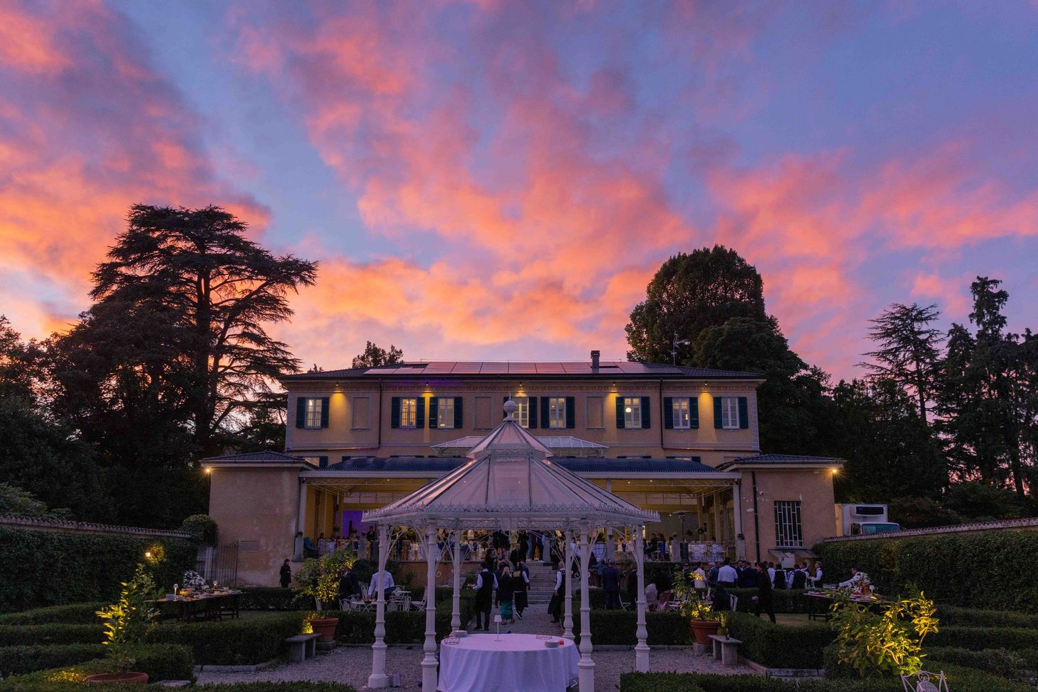 Elegant mansion lit up at dusk with a decorated gazebo and dramatic purple sunset sky.
