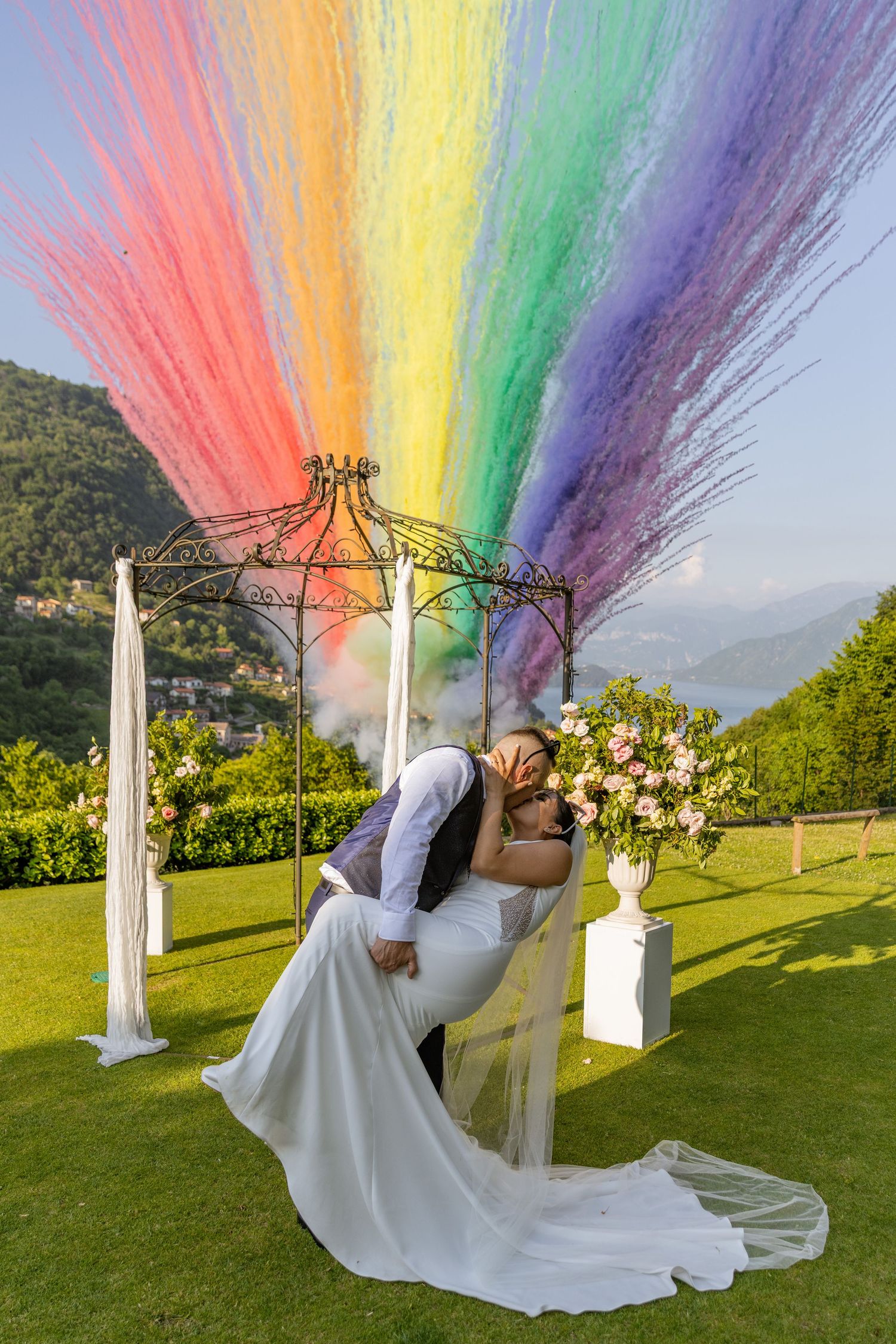 A wedding couple kisses under a vibrant rainbow smoke display on a hilltop garden setting.
