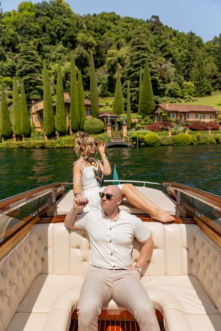 Couple relaxing on a wooden boat with Italian cypress trees and lakeside villa scenery.