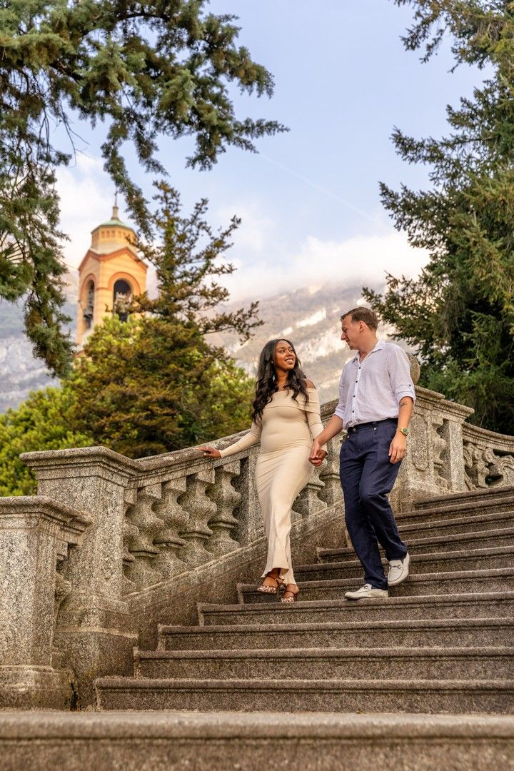 Couple holding hands walking down ornate stone stairs with a church tower and mountains in the background.