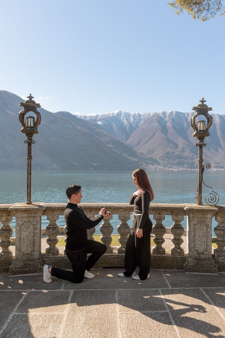 Man proposes on one knee to woman on scenic lakeside terrace with mountain backdrop.