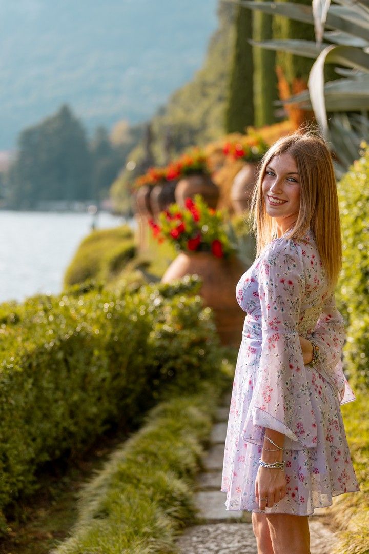 Woman in floral dress poses along scenic lakeside garden path at sunset.