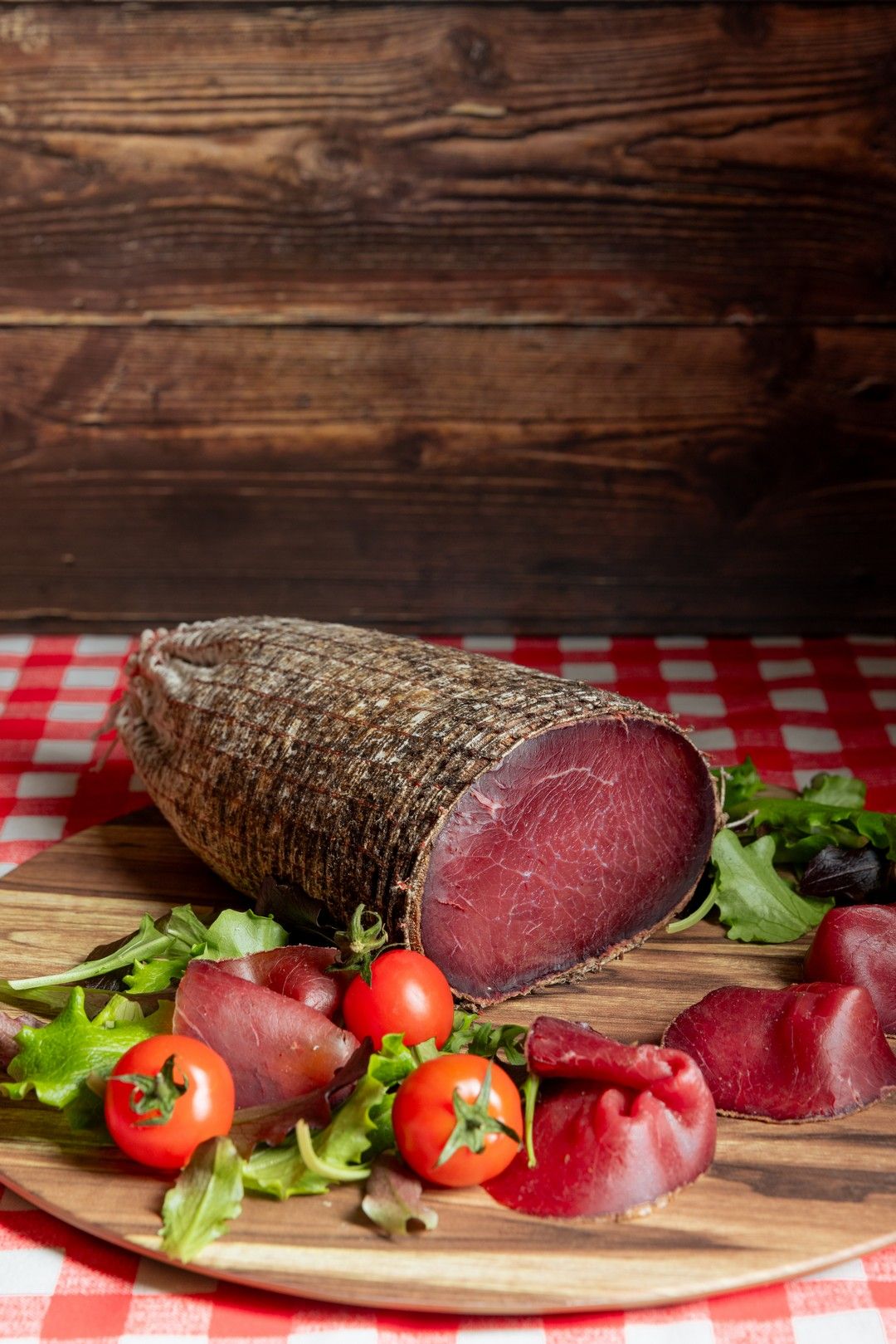 Sliced cured meat with fresh vegetables and herbs on wooden serving board.