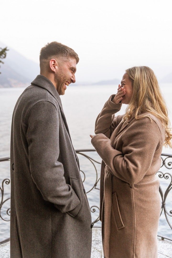 Happy couple in wool coats laughing together on a lakeside promenade on a cloudy day.