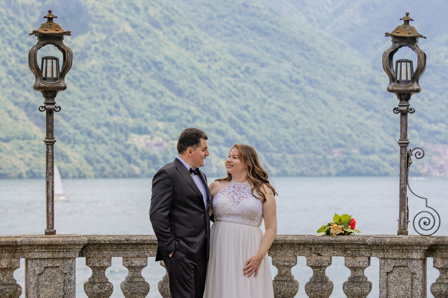 Elegant wedding couple poses along ornate lakeside balustrade with mountain views.