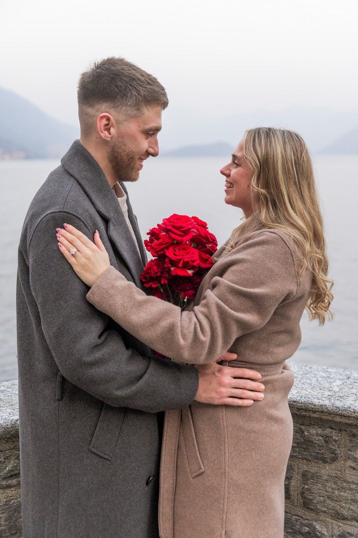 Couple embracing outdoors by a lake, holding red roses bouquet, wearing gray and beige coats, smiling at each other.