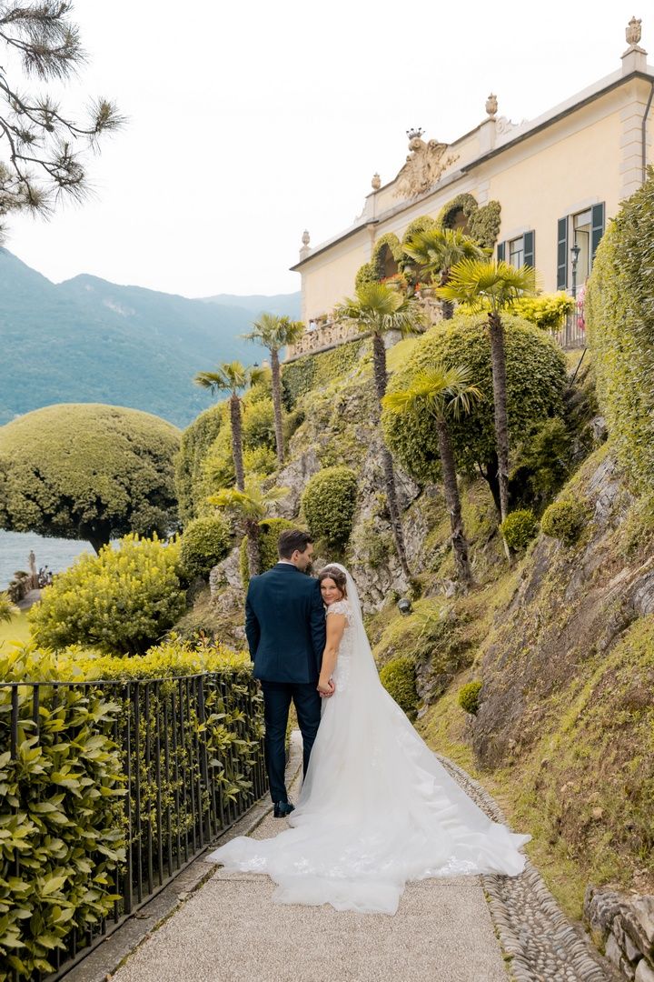 Wedding couple walking along stone path beside elegant villa overlooking mountain lake.