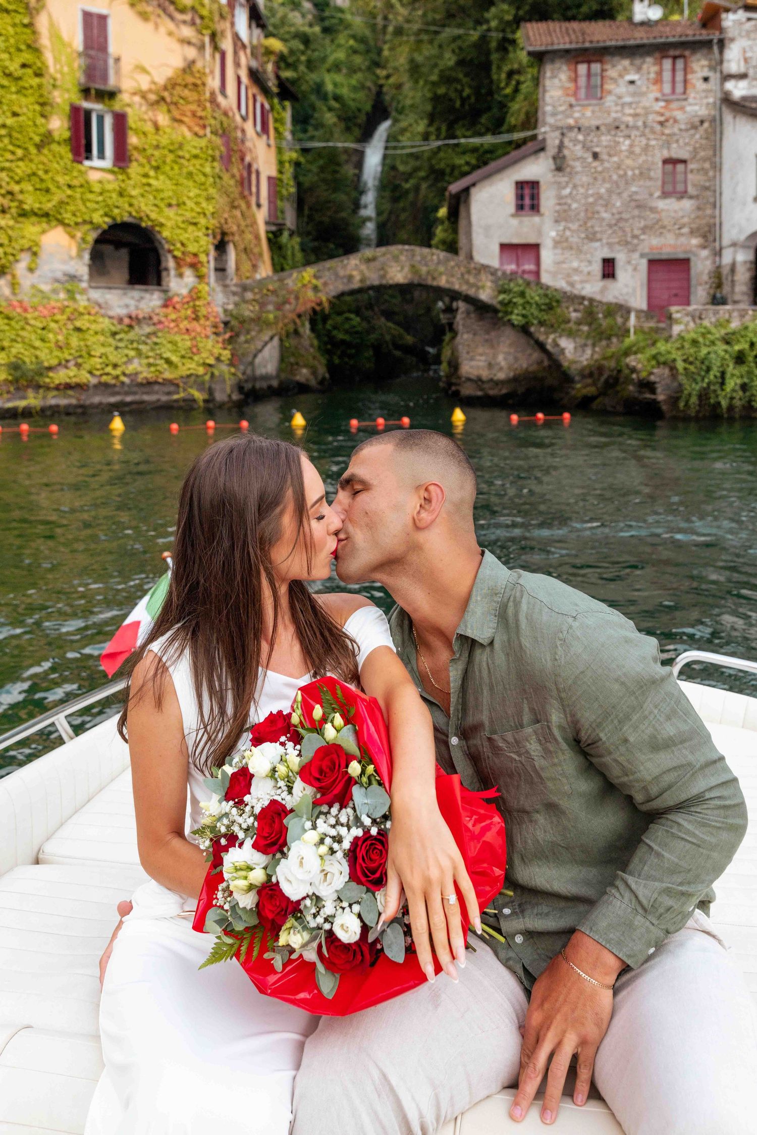 A romantic moment captured on a boat with red roses and stone buildings along the shoreline.