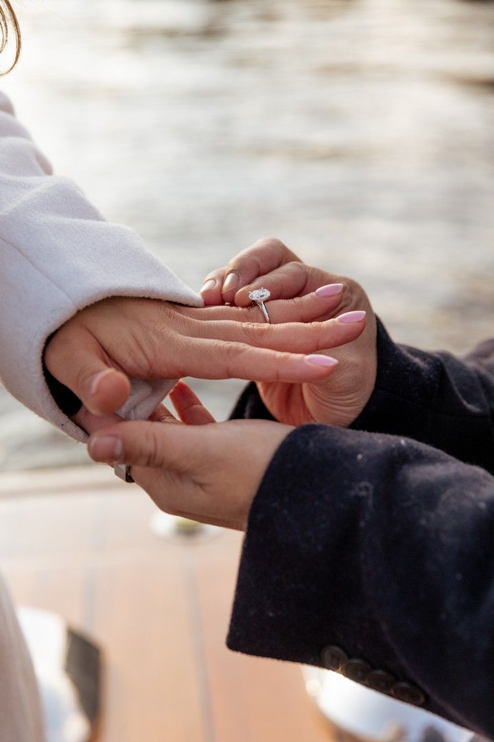 A person placing a diamond engagement ring on their partner's finger outdoors near water.