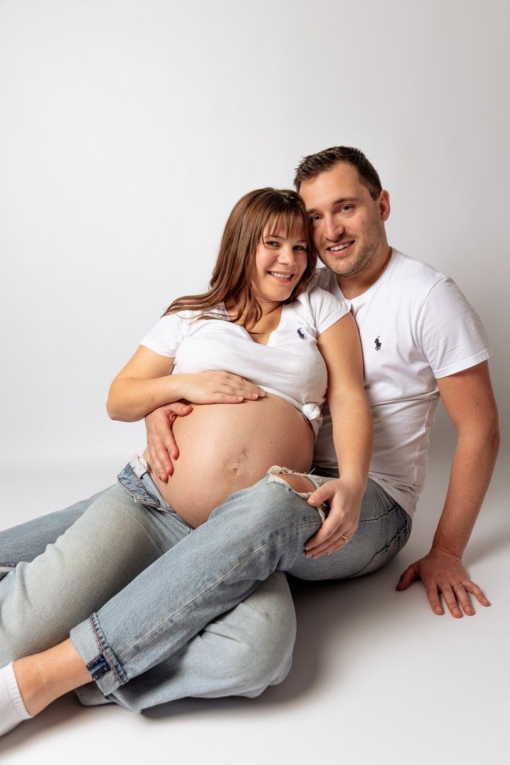 Smiling pregnant couple in white shirts and jeans sitting on floor, man embracing woman while she holds her baby bump.