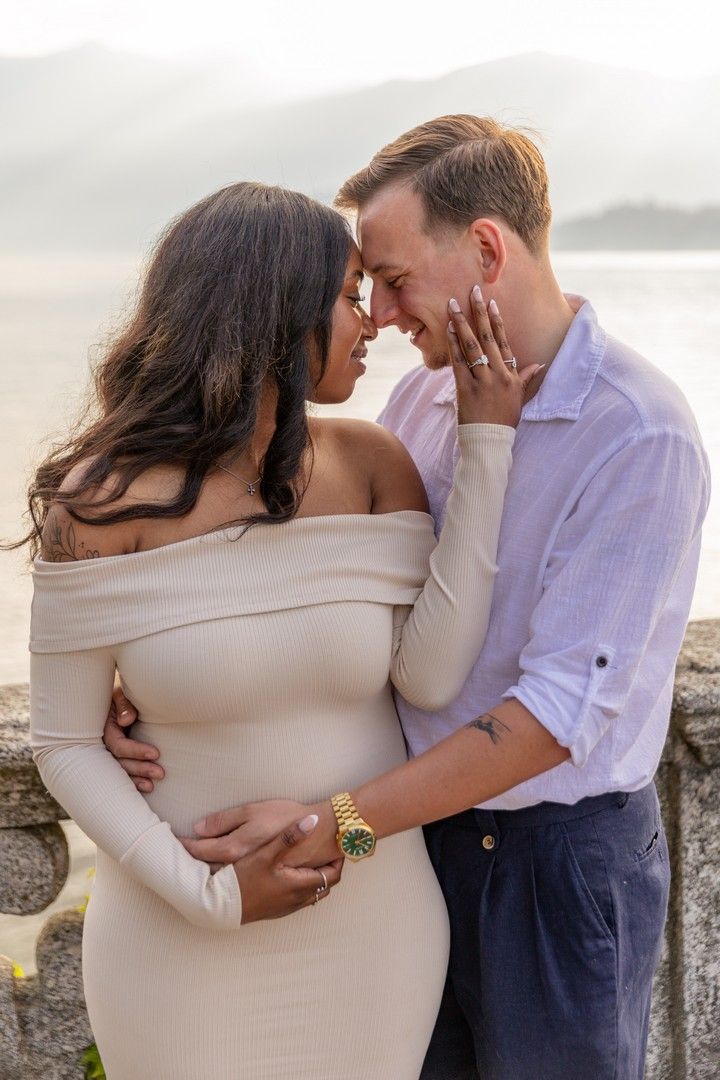 Romantic couple in an intimate embrace outdoors, woman in cream off-shoulder dress, man in white shirt, lakeside backdrop.