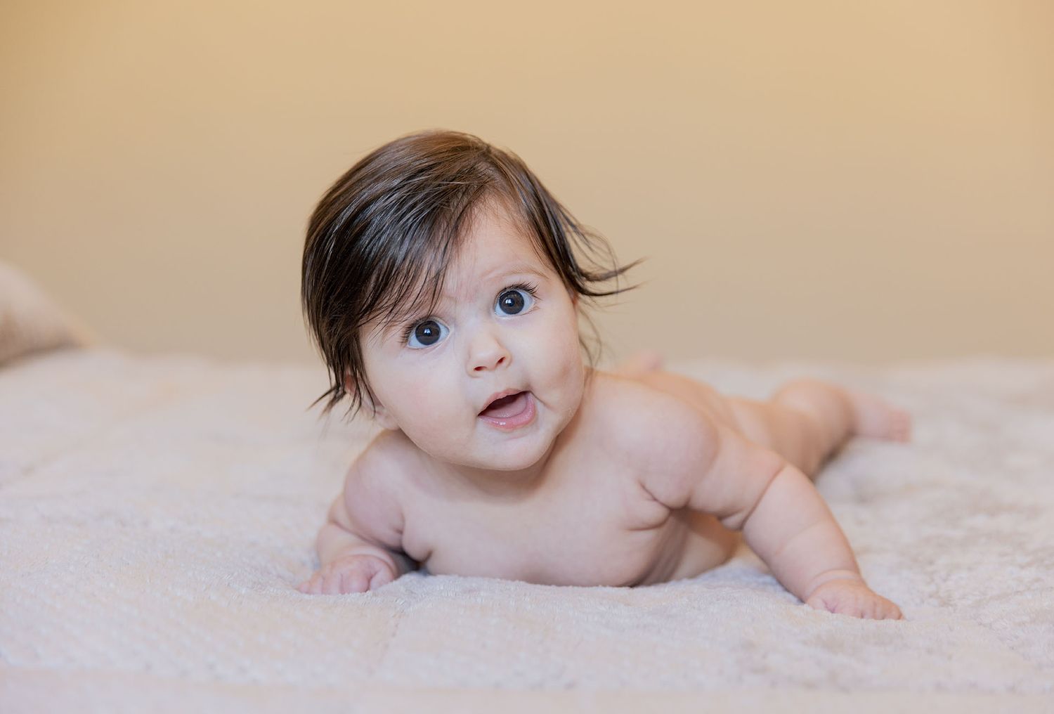 Baby lying on stomach on beige blanket looking up with curious expression.