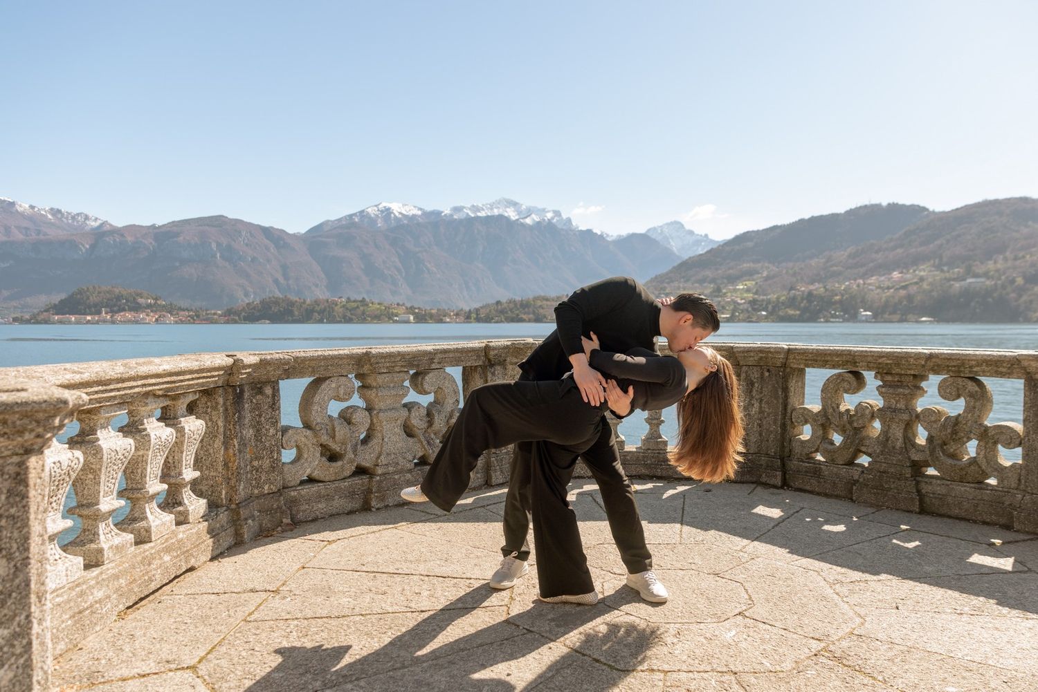 Couple sharing a romantic dip kiss on a stone terrace overlooking a serene alpine lake and mountains.