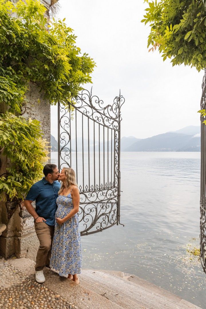 Couple poses by decorative iron gates with lake and mountain landscape in background.