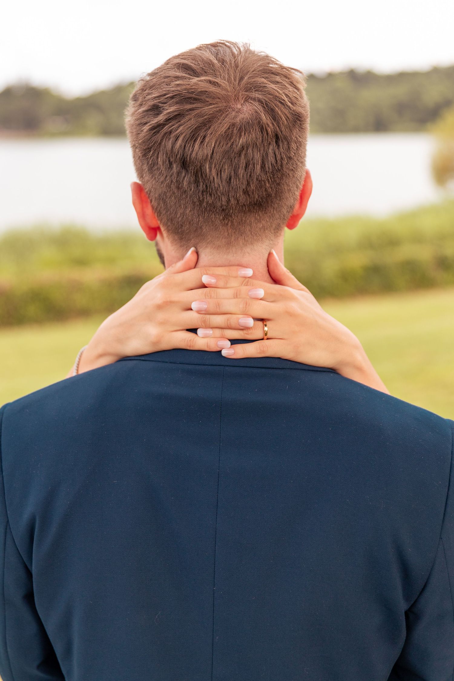 Back view of person in navy suit jacket massaging their neck while standing outdoors near a lake.