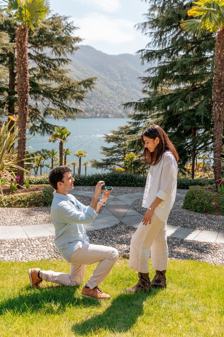 Man proposing on one knee to woman in garden with lake and mountains backdrop on sunny day.