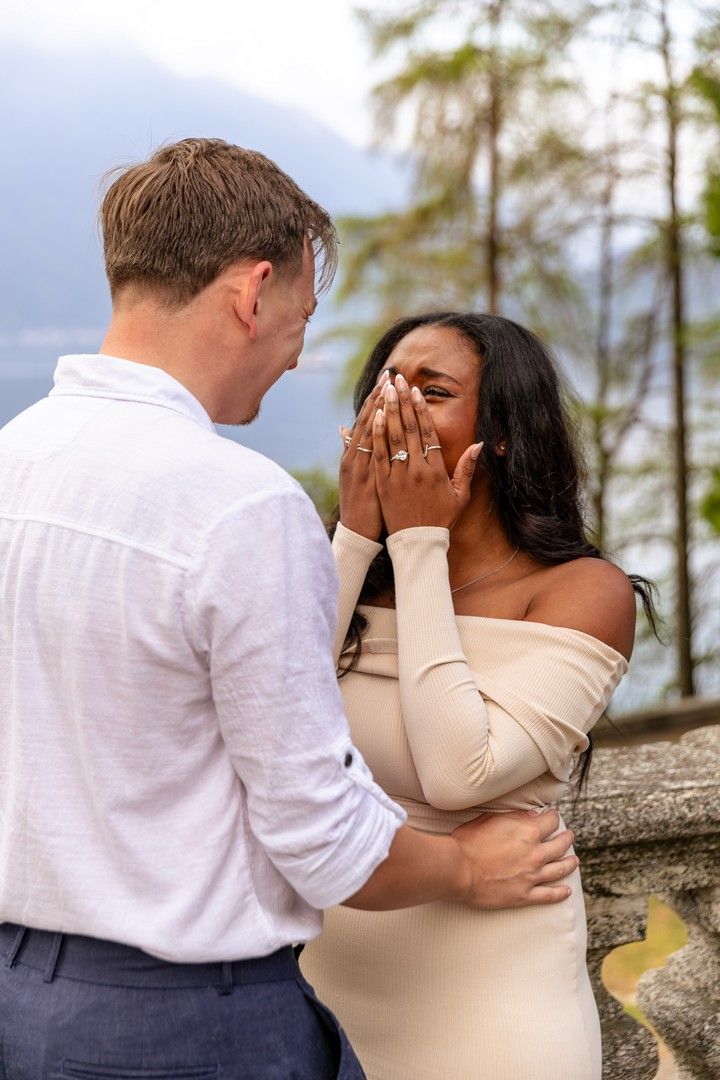 An emotional woman covers her mouth in tears during a marriage proposal outdoors near a lake and mountains.