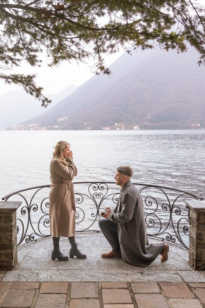 Man kneeling on one knee proposing to a woman by a scenic mountain lake with ornate iron railing.