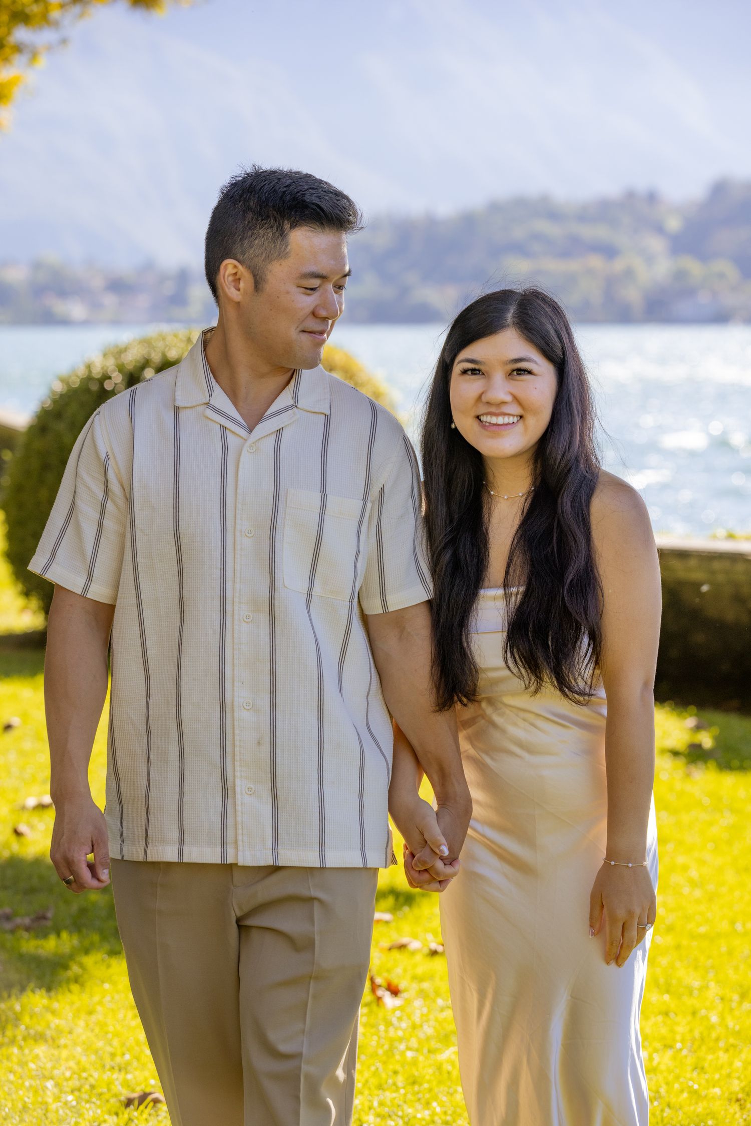A couple walks hand in hand through a sunny meadow with water views in the background.